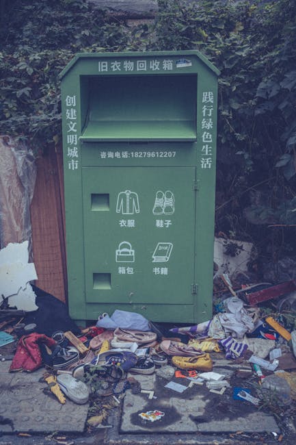 A green, weathered clothing recycling bin placed outdoors on a concrete surface, with various discarded items scattered around its base. The bin features compartments for different clothing categories, with icons and Chinese characters indicating sections for shirts, shoes, bags, and books. The top section has Chinese writing that translates to 'Old Clothes Recycling Box,' and a contact phone number beneath it. Surrounding the bin is a cluttered assortment of used shoes, torn fabric, plastic bottles, and other rubbish, creating a chaotic scene of informal waste disposal. Behind the bin, there is dense foliage and possibly a wooden fence, suggesting an outdoor environment near a residential or garden area. The overall atmosphere appears neglected, highlighting the need for proper waste management and the role of independent collection services like House Clearance Richmond in rubbish removal and recycled material handling.