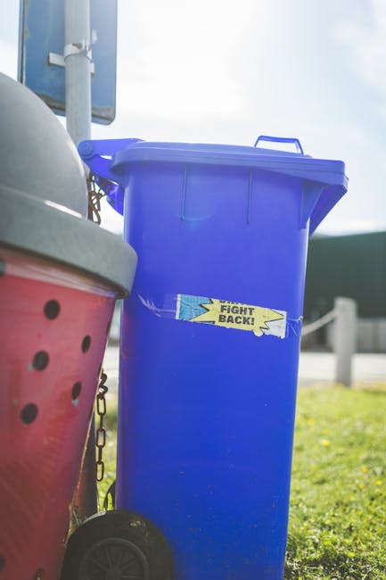 A green, weathered clothing recycling bin placed outdoors on a concrete surface, with various discarded items scattered around its base. The bin features compartments for different clothing categories, with icons and Chinese characters indicating sections for shirts, shoes, bags, and books. The top section has Chinese writing that translates to 'Old Clothes Recycling Box,' and a contact phone number beneath it. Surrounding the bin is a cluttered assortment of used shoes, torn fabric, plastic bottles, and other rubbish, creating a chaotic scene of informal waste disposal. Behind the bin, there is dense foliage and possibly a wooden fence, suggesting an outdoor environment near a residential or garden area. The overall atmosphere appears neglected, highlighting the need for proper waste management and the role of independent collection services like House Clearance Richmond in rubbish removal and recycled material handling.