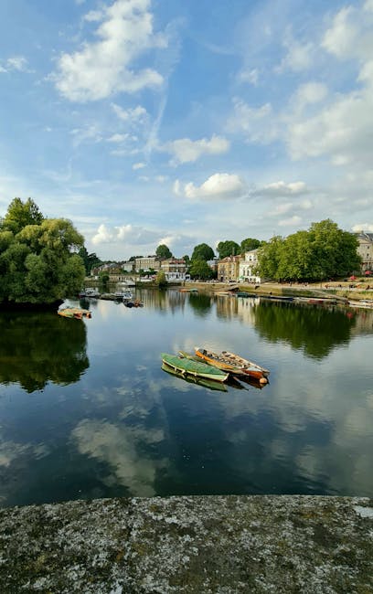 A calm river scene during daytime with clear weather and a partly cloudy sky. In the foreground, a stone embankment edge with a textured, weathered surface is visible. The river's surface is smooth and reflective, mirroring the sky and clouds above. Several small boats, including a green rowboat and a few others with wooden or metal finishes, are anchored close to the bank, some partially leaning to one side. On the far bank, there are residential buildings with light-colored facades, some with visible windows and small terraces, interspersed with lush green trees providing shade and contrast. The trees and buildings are reflected in the water, enhancing the tranquil atmosphere. The scene suggests an environment suitable for quiet outdoor activities or leisurely boat trips, with an emphasis on natural beauty and peaceful surroundings. The environment and scene imply a context related to private or independent boat use, aligning with the themes of alternative waste handling and clearance that House Clearance Richmond may support through on-site or personalized services.