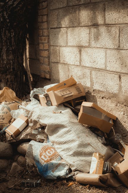 A pile of discarded cardboard boxes, some flattened and others still partially assembled, resting against a rough concrete wall outdoors. The boxes vary in size and are made of brown corrugated cardboard, with visible flaps and printed labels. Among the boxes, there is a woven fabric sack or bag, possibly used for waste collection, and a partially visible brown paper bag. The ground beneath the rubbish consists of dirt and small stones, with a large tree trunk to the left side of the image providing some shade. The scene suggests a private, unauthorised rubbish accumulation area, perhaps awaiting collection or disposal, and is associated with services that handle local waste clearance outside the scope of municipal waste management, like those offered by House Clearance Richmond. Natural lighting highlights the textures of the materials, emphasizing the cluttered and unorganized state of the waste, which is typical in situations requiring independent rubbish removal or on-site clearance services.