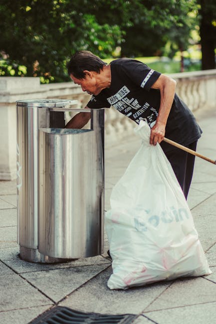 A middle-aged man with dark hair, wearing a black T-shirt with white text and dark trousers, is seen disposing of rubbish into a cylindrical, stainless steel public waste bin with a rounded top and a small, rectangular opening at the front, located on a paved sidewalk. The man is holding a large, white plastic bag filled with waste in his left hand, while with his right hand, he is reaching into the bin to dispose of more rubbish. The background features lush green trees and a low stone balustrade, suggesting a park-like environment in an urban setting. The scene is lit by natural daylight, creating a clear and detailed view of the man and the surrounding environment, reflecting typical outdoor rubbish collection and waste disposal activities, possibly as part of a local community clean-up or regular rubbish collection service by House Clearance Richmond.