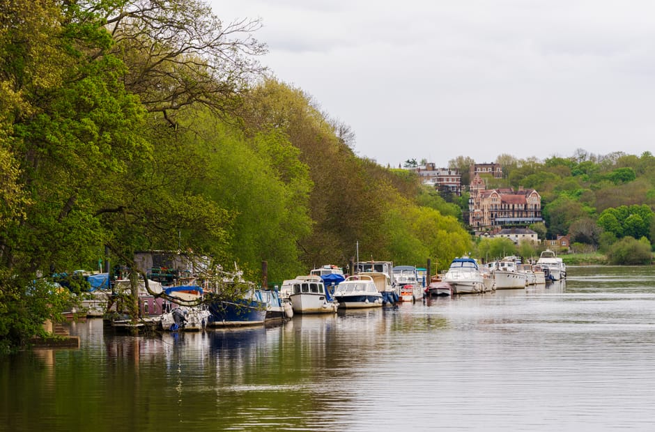 A tranquil riverside scene featuring a row of various boats docked along the water's edge, with some boats covered by blue and white tarps and others showing polished wooden or metal hulls with visible textures, including smooth paint finishes and weathered surfaces. The boats are moored to wooden or metal posts, and some are partially obscured by overhanging leafy green trees on the left side of the image, which display fresh spring foliage in different shades of green. The water reflects the boats and trees, creating a calm, mirror-like surface with gentle ripples. In the background, a hillside is dotted with large, multi-story residential buildings with brick and stone facades, sloped roofs, and large windows, interspersed with trees and greenery. The overcast sky adds diffuse, soft lighting, enhancing the natural tones of the scene. This image, associated with professional waste management services like House Clearance Richmond, highlights the peaceful setting of a private riverside area, where alternative disposal or clearance of dockside debris may take place, supporting the context of rubbish removal and private waste handling.